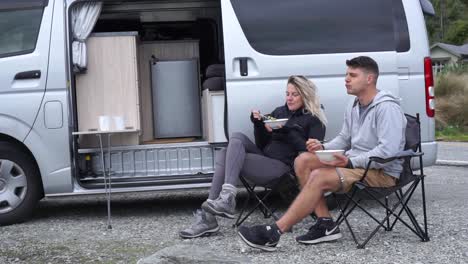 SLOWMO---Young-attractive-couple-sitting-and-eating-lunch-with-motorhome-by-Lake-Wakatipu,-Queenstown,-New-Zealand