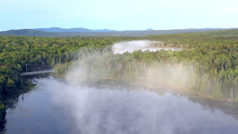 Aerial-drone-shot-rising-above-the-mist-and-still-blue-waters-of-the-Spectacle-Ponds-and-a-country-road-with-cars-winding-through-the-green-forests-of-the-Maine-wilderness