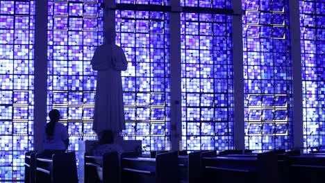 Totale-Aufnahme-Zweier-Menschen,-Die-Vor-Einer-Skulptur-In-Der-Dom-Bosco-Kirche-Beten,-Die-Durch-Die-Blauen-Fenster-Beleuchtet-Wird
