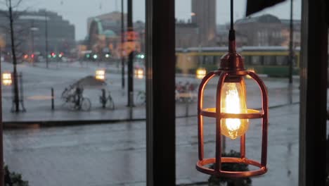 lantern-lightbulb-revealing-behind-leaves-shadows,-with-rainy-city-in-background-and-man-passing-with-umbrella