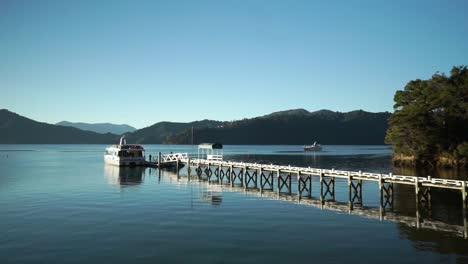 Cruise-tourist-tour-boat-at-dock-by-luxury-lodge-in-Marlborough-Sounds,-New-Zealand-with-small-boat-and-mountains-in-background-during-early-morning