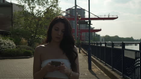Attractive-hispanic-latina-woman-checking-her-phone-while-walking-towards-the-camera,-during-a-bright-day-at-the-banks-of-the-river-Thames-in-London-United-Kingdom