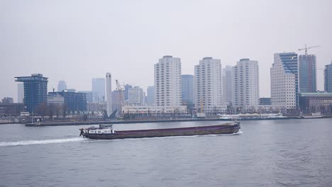 Large-cargo-ship-passing-through-the-city-of-Rotterdam-with-skyscrapers-of-the-financial-district-partly-under-construction-in-the-background-on-a-rainy-overcast-day