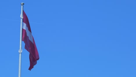 Große-Lettische-Flagge-Weht-An-Sonnigen-Tagen-Langsam-Am-Blauen-Himmel,-Flugzeug-Fliegt-Durch-Den-Hintergrund,-Mittlere-Aufnahme