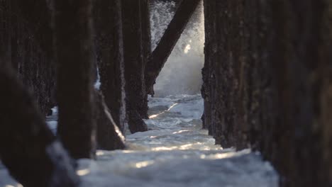 Waves-crashing-under-a-pier,-old-wooden-pillings-with-sunbeams-coming-through
