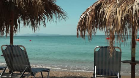 Griechischer-Sandstrand-Mit-Liegestühlen-Und-Palmenschirmen,-Während-Kristallklares-Und-Hellblaues-Wasser-Auf-Die-Küste-Trifft