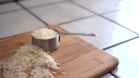 Ein-Stapel-Weißer-Reiskörner-Und-Ein-Messbecher-Für-Zutaten-Auf-Einem-Holzschneidebrett-In-Einer-Küchenrutsche-Nach-Links