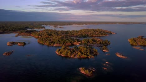 Kleine-Felsige-Inseln-Mit-Grünen-Kiefern-Im-Klaren-Blauen-See-Bei-Sonnenuntergang,-Drohnen-Luftweite-Umlaufbahn