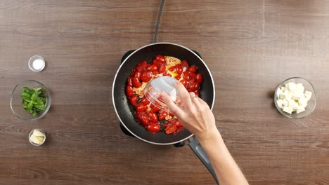 Top-down-shot-of-pouring-milk-from-glass-bowl-into-large-pan-with-sauteed-cherry-tomatoes-placed-on-stove-on-wooden-table-with-other-ingredients-around
