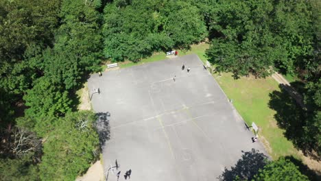 An-aerial-view-over-people-playing-basketball-on-a-court-is-surrounded-by-beautiful-green-trees