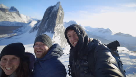 A-Group-of-Three-Hikers-Smile-and-Laugh-at-the-Camera-from-on-Top-of-a-Snowy-Mountain-Peak-with-Segla-in-the-Background