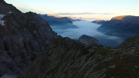 Drone-shot-flying-forwards-towards-Gleckstein-hut-building-that-stands-on-the-very-edge-of-a-steep-and-high-cliff-of-the-mountain-top-in-the-alps-of-Switzerland-during-a-quiet-and-cold-sunrise