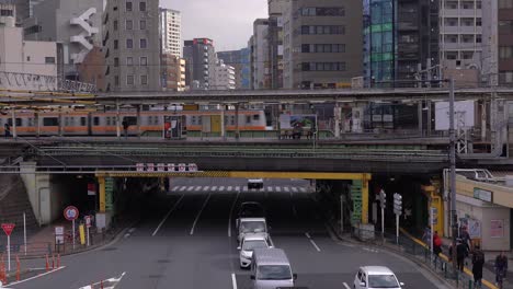 Train-Passing-Over-Elevated-Rail-Tracks-In-Tokyo-Japan-With-Light-Traffic-On-The-Road-Under-And-High-Rise-Buildings-In-The-Background---wide-shot