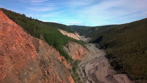 Exhilarating-wilderness-flight-hugging-mountainside-and-red-bedrock-by-side-of-cliff-edge-in-Yukon-Burwash-creek-towards-sensational-majestic-blue-sunny-sky-day,-Canada,-aerial-approach