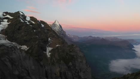 Slow-and-gentle-aerial-shot-flying-next-to-the-peak-of-the-alpine-mountains-in-Switzerland-during-a-sunrise-with-red,-orange-and-pink-colors-in-the-sky