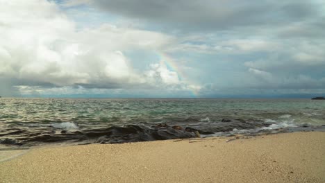 Waves-breaking-on-rocky-shore-of-tropical-island-with-rainbow-and-clouds-as-backdrop