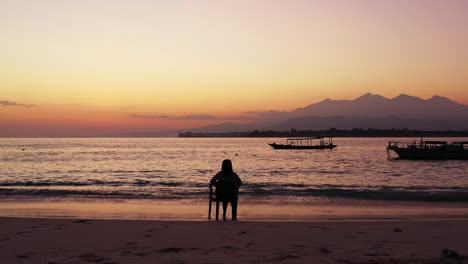 Silhouette-of-girl-sitting-on-chair-over-sandy-beach,-watching-colorful-sky-after-sunset-with-mountains-horizon-and-anchored-boats-on-calm-lagoon,-Bali