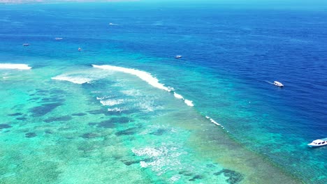 Boote-Segeln-über-Das-Blaue-Meer-Entlang-Der-Türkisfarbenen,-Flachen-Lagune-Mit-Wunderschönem-Meeresboden-Voller-Korallenriffe-Und-Felsen-In-Antigua
