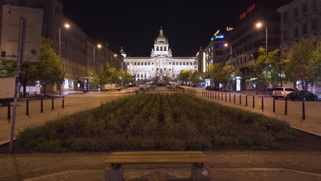 A-view-of-the-National-museum-in-Prague,-Czechia-on-the-empty-Wenceslas-square-in-the-historical-centre,-lit-by-street-lamps-during-a-Covid-19-lockdown,-pedestal-shot-over-a-bench-and-a-flower-patch