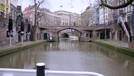 Canal-boat-slowly-cruising-along-canal-in-Utrecht,-Netherlands,-underneath-a-brickwork-bridge,-with-a-Netherlands-and-European-Union-flag-flying-in-the-background