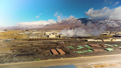 Industrial-complex-emits-smoke-and-steam-in-an-urban-valley-under-the-mountains---aerial-view-with-orbiting,-parallax-motion