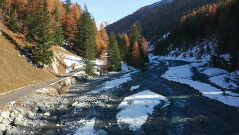 forward-flight-over-a-small-river-at-the-Swiss-National-Park,-with-some-snow-on-the-ground-and-beautiful-autumn-coloured-trees