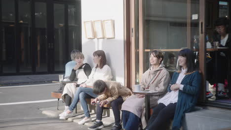 Japanese-People-Happy-Talking,-Sitting-And-Leaning-On-Wall-While-Drinking-Coffee-Outside-A-Cafe-Restaurant-In-Street-Of-Tokyo,-Japan