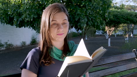 Young-caucasian-student-woman-slowly-turns-page-of-book-outdoors