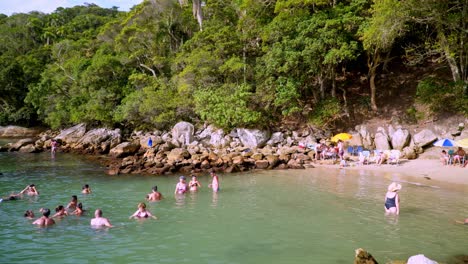 Pan-right-of-people-having-fun-in-the-sea-near-a-rocky-shore-and-woods-in-Praia-da-Sepultura-beach,-Brazil