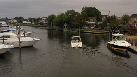 A-low-angle,-aerial-view-of-a-fishing-boat-sailing-through-a-canal-behind-houses-on-Long-Island-on-a-cloudy-day
