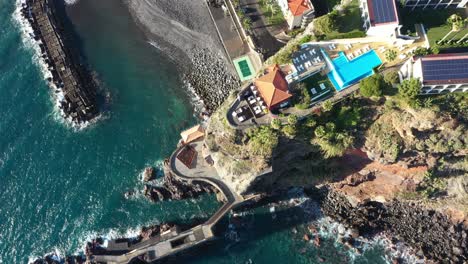 Drone-shot-moving-upwards-slowly,-looking-down-on-a-luxury-resort-sitting-on-top-of-the-cliff-on-the-coast-of-Madeira-with-the-ocean-during-a-sunset