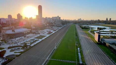 Slow-aerial-dolly-in-shot-along-horse-racing-track-at-famous-racecourse-Hipodromo-Argentino-de-Palermo-in-capital-city-Buenos-Aires,-Argentina,-with-cityscape-and-sunset-in-the-background