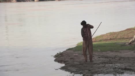 Environmental-Portrait,-A-Pakistani-Man-Flicks-Mud-Into-A-River-With-A-Walking-Stick
