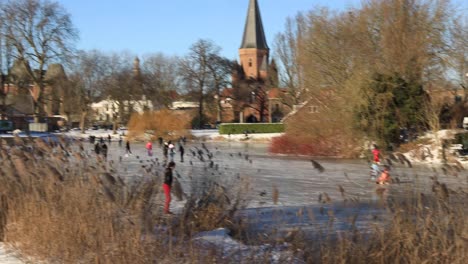 Left-pan-showing-ice-skating-people-on-the-canal-with-reed-in-the-foreground-revealing-historic-medieval-Walburgiskerk-and-Drogenapstoren-rising-up-in-the-background