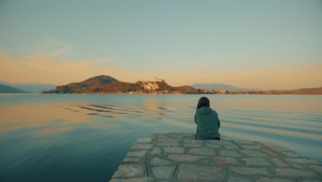 Back-view-of-woman-with-green-hoodie-sitting-on-jetty-edge-looks-at-Angera-castle-overlooking-Maggiore-lake-in-Italy