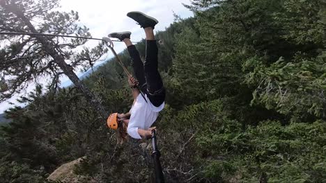 Vertical-action-camera-shot-of-a-young-man-hanging-upside-down-in-a-zipline-in-Canada-with-big-trees-around-and-under-him
