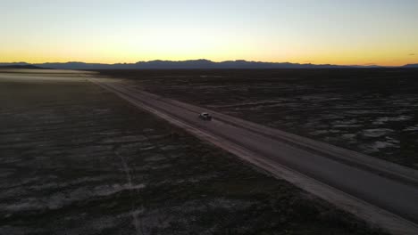 Truck-driving-down-a-dusty-dirt-road-in-a-desert-basin-at-sunset---aerial-view-showing-the-landscape-and-mountains-in-the-distance