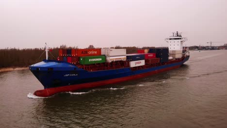 Shortsea-container-vessel-JSP-Carla-loaded-with-containers-on-deck-sailing-on-high-speed-over-the-Dutch-river-Oude-Maas-towards-the-cargo-port-Moerdijk-on-a-cloudy-day