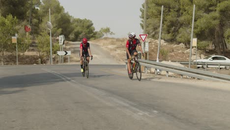 Dos-Profesionales-Ciclistas-De-Carretera-En-Cámara-Lenta-Montando-En-La-Carretera-Vacía-Del-Bosque-Pidiendo-Perdón-A-Los-Conductores-Detrás-De-Ellos