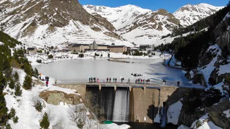 Aerial:-dam-in-a-frozen-lake-and-people-walking-along,-among-mountains-and-a-sharine-in-the-background