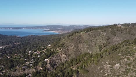 Small-town-on-forestry-hillside-with-beautiful-blue-bay-in-horizon,-high-angle-drone-view