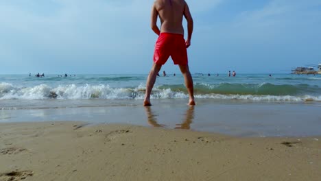 Low-angle-ground-surface-level-pov-of-boy-kicking-red-ball-from-beach-to-sea-water-with-man-jumping-to-save