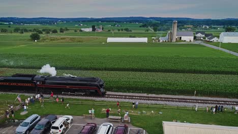 Eine-Parallele-Luftaufnahme-Eines-Antiken-Dampf-Personenzuges,-Der-Rauch-Ausbläst-Und-An-Familien-Und-Kindern-Vorbeifährt,-Die-Ihm-Dabei-Zusehen,-Wie-Er-Durch-Das-Farmland-Von-Pennsylvania-Fährt