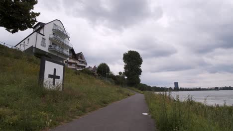 Cologne,-North-Rhine-Westphalia,-Germany,-circa-2019:-Establisher-shot-of-a-bike-lane-along-the-Rhine-south-of-the-City-of-Cologne-with-a-big-white-cross-sign-for-ships-along-the-river