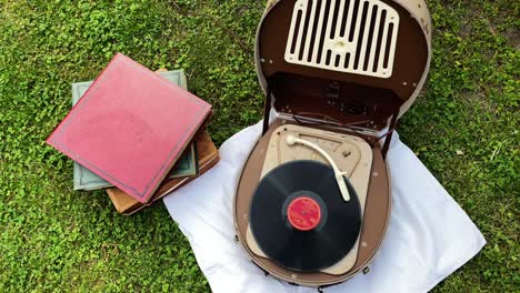 Top-view-of-an-old-retro-record-player-from-the-1950s-and-1960s-is-used-outside-in-the-garden-to-play-music-from-an-old-vinyl-record-and-next-to-it-is-a-stack-of-old-vinyl-records
