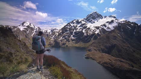 Pan,-hiker-overlooks-alpine-lake-in-snow-capped-mountain-landscape,-Routeburn-Track-New-Zealand