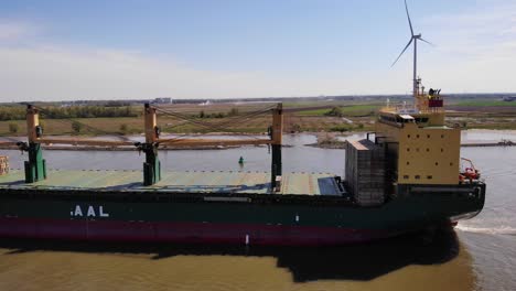 Aerial-Tracking-Shot-Of-Port-Side-View-Of-Aal-Paris-Cargo-Ship-Along-Oude-Maas-With-Still-Wind-Turbine-In-Background