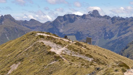 Static,-distant-hiker-approaches-remote-alpine-shelter,-surrounding-mountains,-Fiordland,-Kepler-Track-New-Zealand