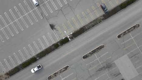 Static-birds-eye-view-of-a-parking-lot-and-mall-access-road-with-traffic-passing-and-lot-light-poles-visible