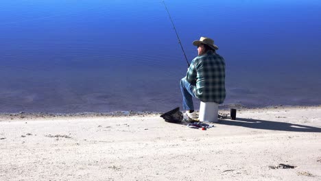 Fisherman-sits-on-an-upside-down-bucket-on-a-beach-and-casts-out-his-fishing-line